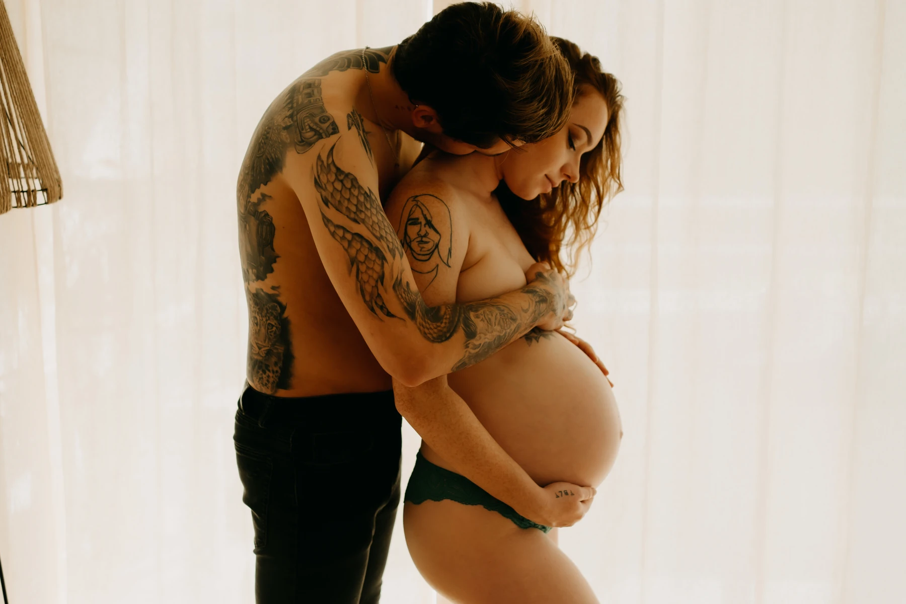 Couple tatoué en attente de bébé, enlacé tendrement lors d'une séance photo grossesse à domicile
