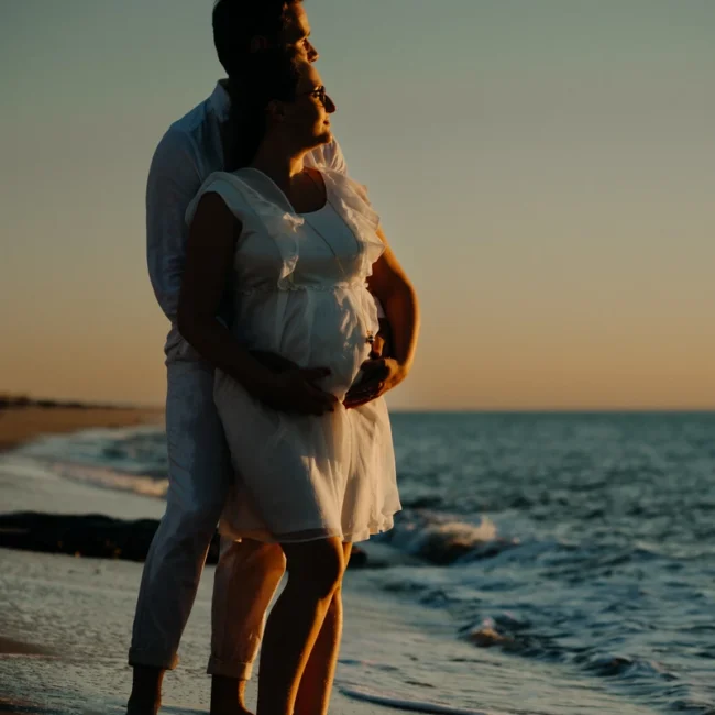 Couple de futurs parents enlacés au bord de l’océan au coucher du soleil