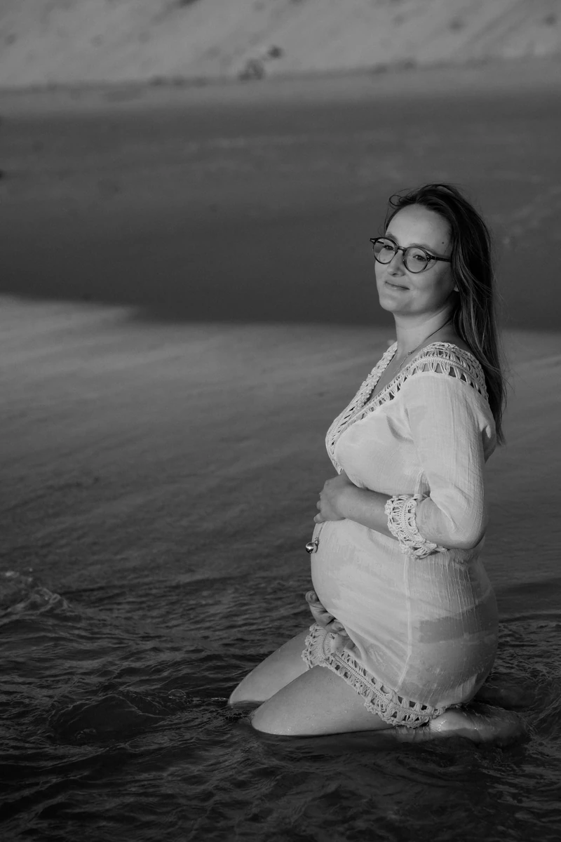 Photo de grossesse sur la plage du Petit Nice au coucher du soleil, future maman à genoux dans l’eau, regard apaisé vers l’objectif