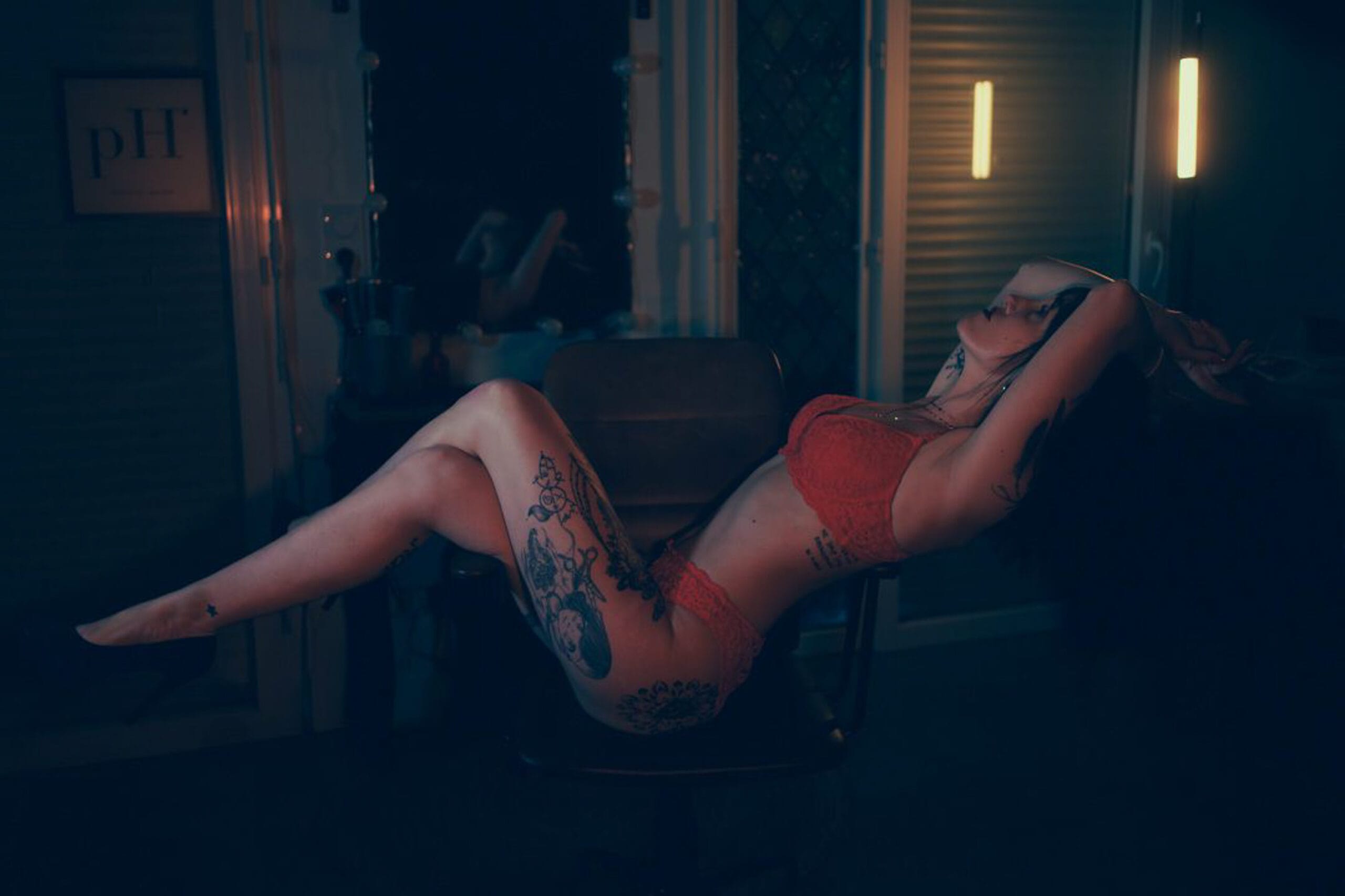 Photo boudoir d’une femme tatouée en lingerie rouge, pose sensuelle sur une chaise, lumière tamisée à Arcachon