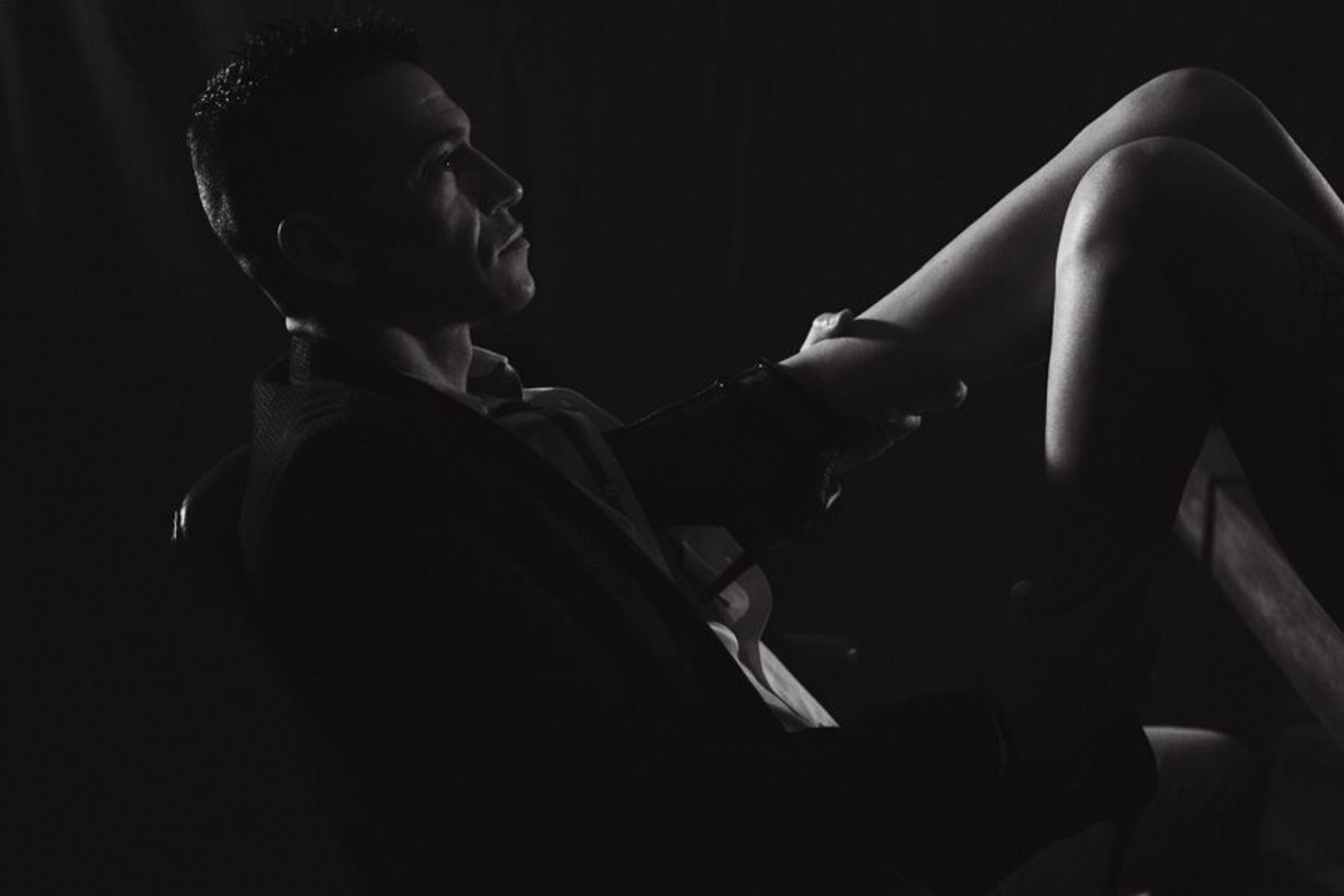 Portrait boudoir artistique en noir et blanc d’un homme en costume caressant la jambe de sa partenaire.