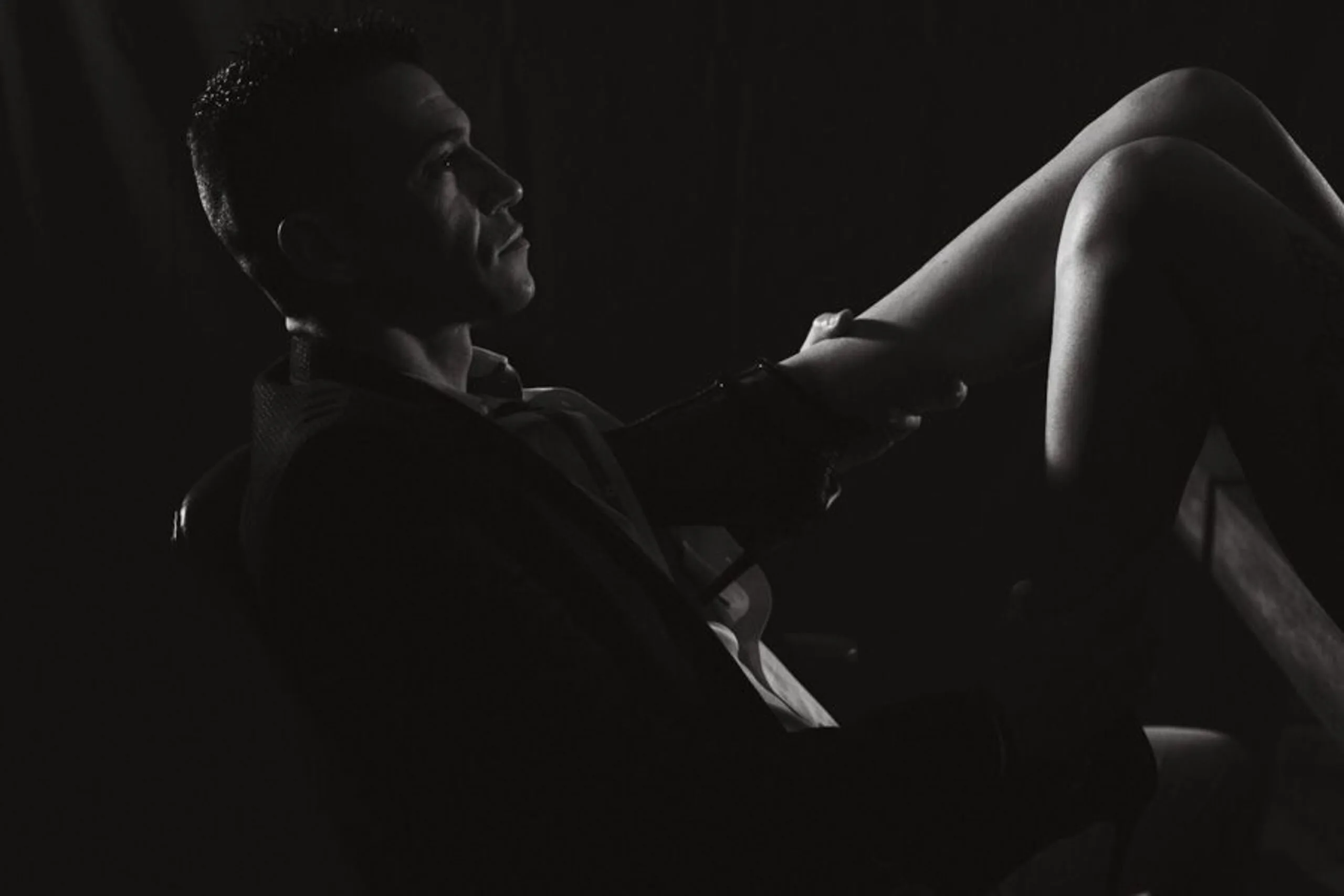 Portrait boudoir artistique en noir et blanc d’un homme en costume caressant la jambe de sa partenaire.