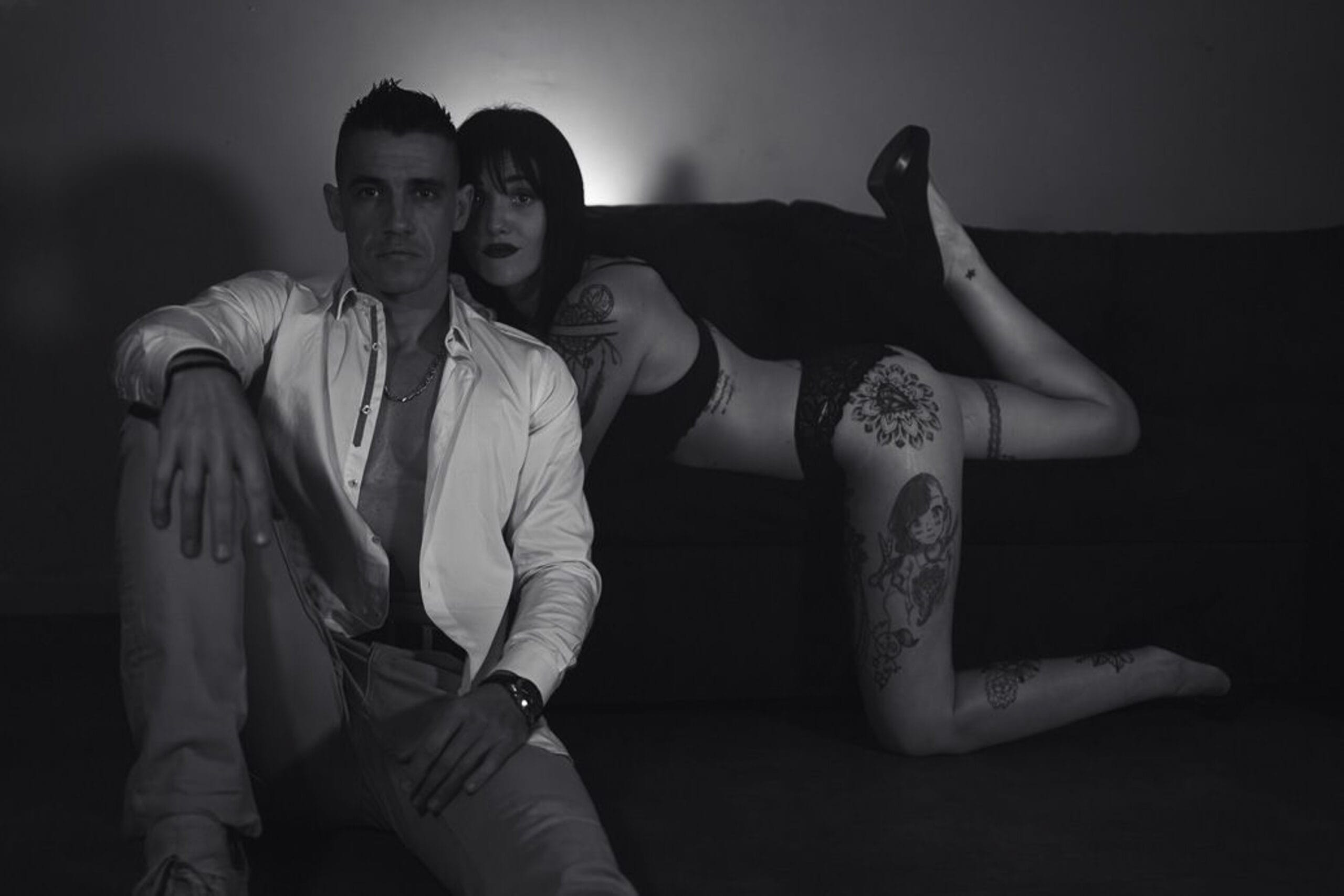 Photo boudoir en noir et blanc d’un couple, assis sur un canapé, avec poses affirmées et jeux d’ombres.
