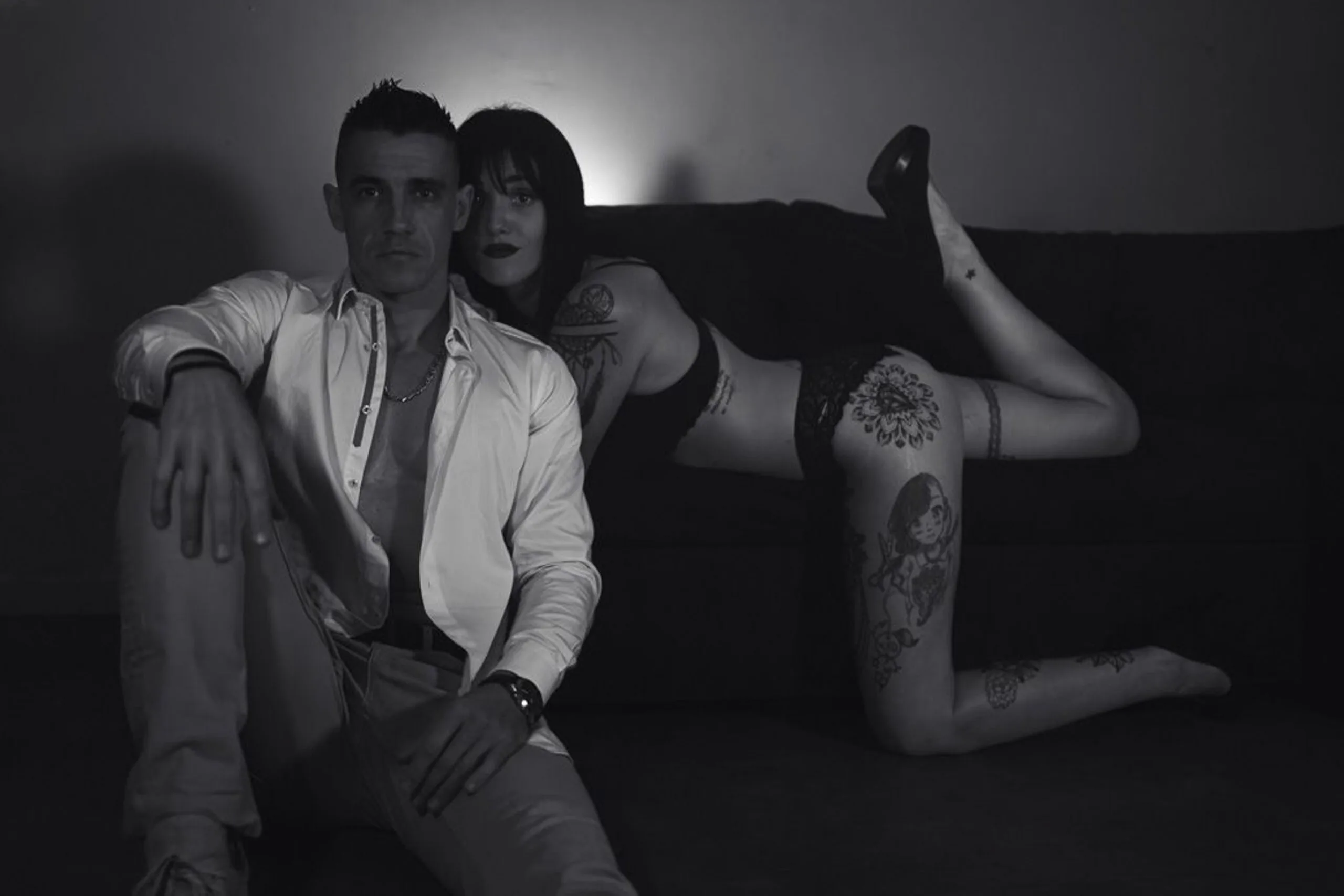 Photo boudoir en noir et blanc d’un couple, assis sur un canapé, avec poses affirmées et jeux d’ombres.