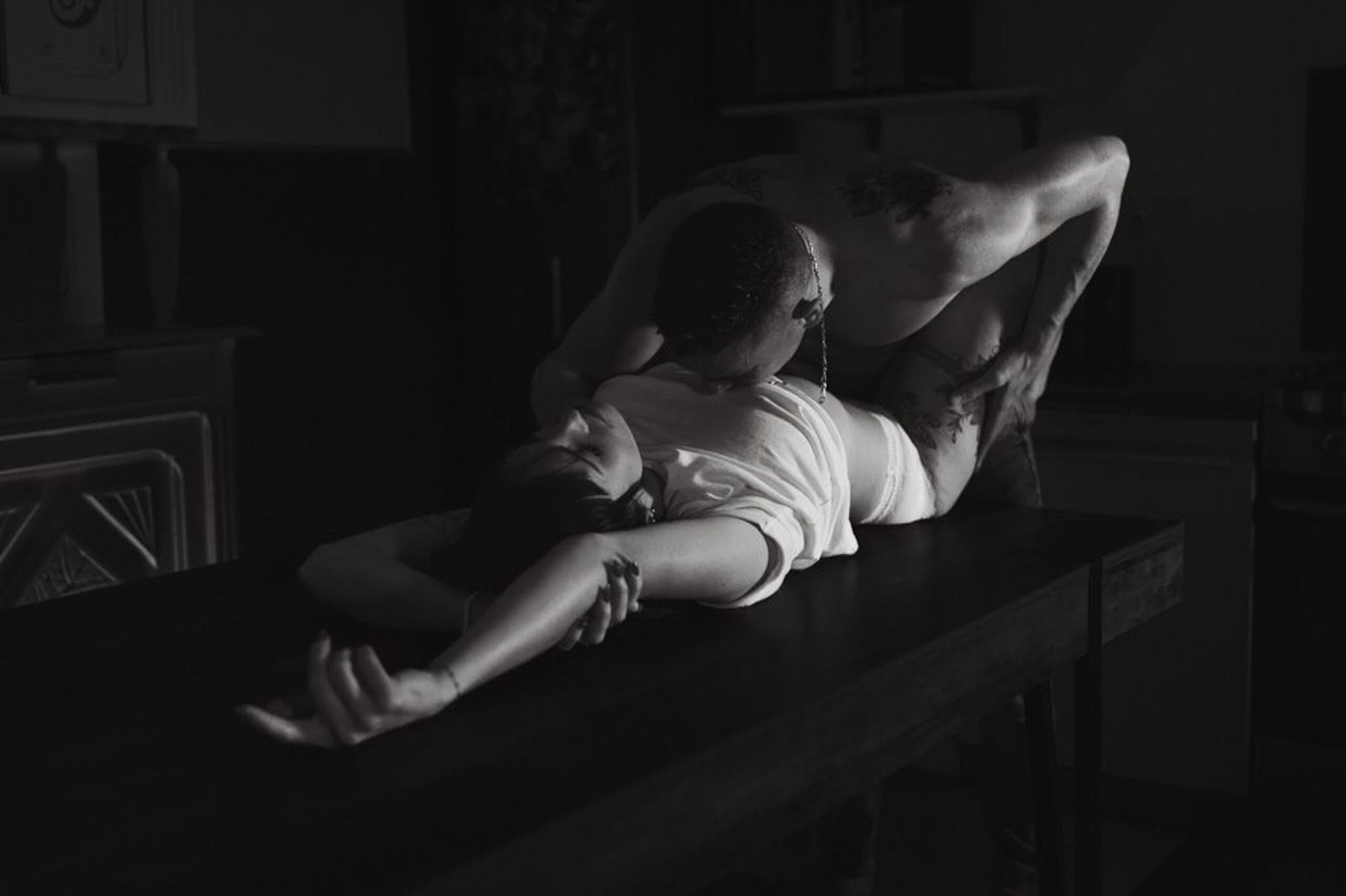 Séance photo boudoir en noir et blanc, couple sur une table, femme allongée bras tendus, lumière dramatique.