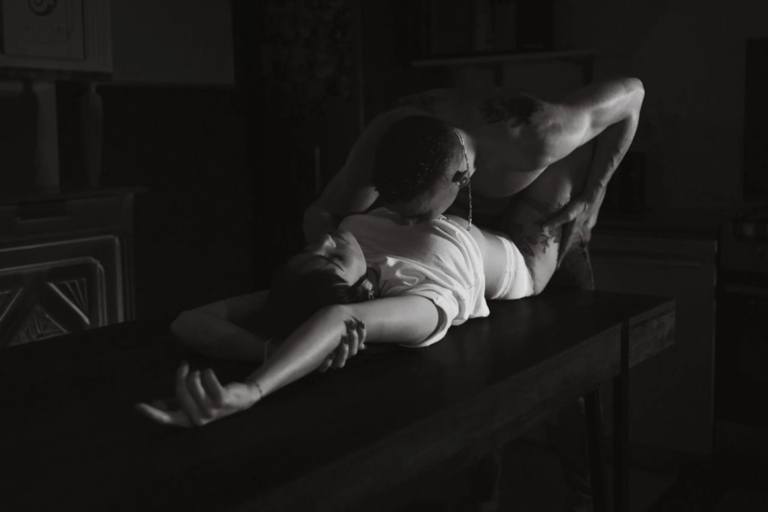 Séance photo boudoir en noir et blanc, couple sur une table, femme allongée bras tendus, lumière dramatique.