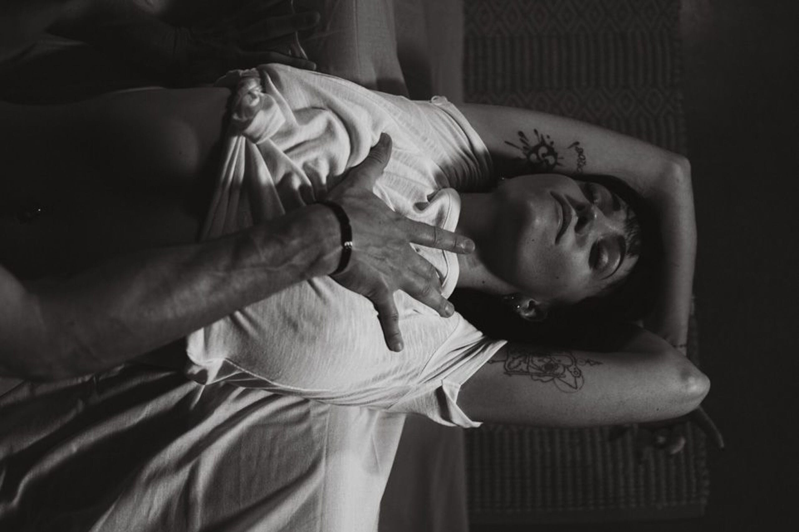 Séance photo boudoir en noir et blanc, femme allongée bras levés, main masculine posée sur sa poitrine.