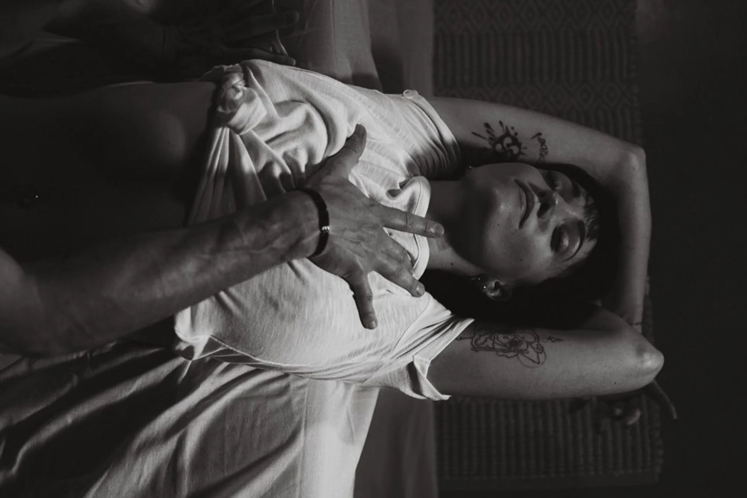 Séance photo boudoir en noir et blanc, femme allongée bras levés, main masculine posée sur sa poitrine.