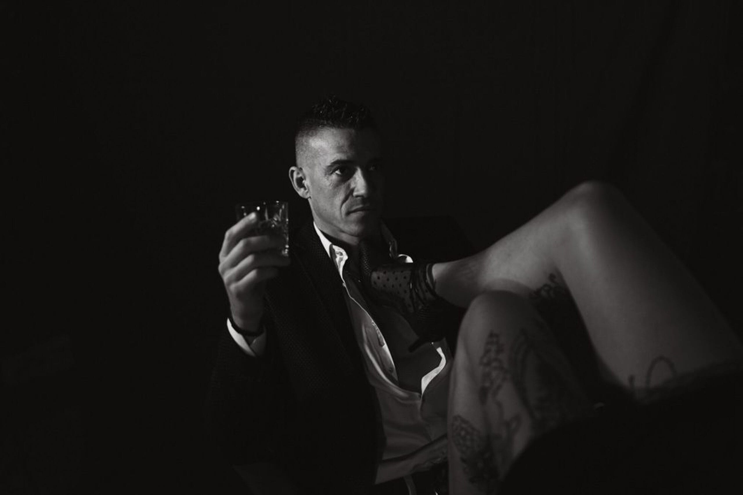 Portrait boudoir noir et blanc d’un homme en costume avec un verre à la main et la jambe tatouée de sa partenaire en avant-plan.